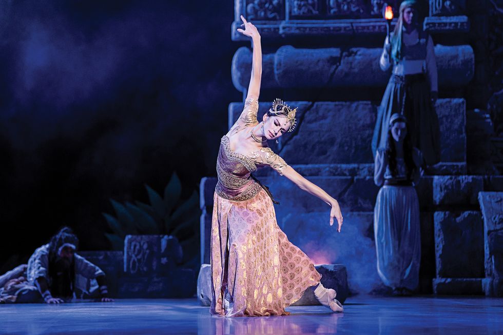 Semionova in a pink and gold outfit and crown, in a deep pli\u00e9 onstage with one leg behind her and her arms framing her face.