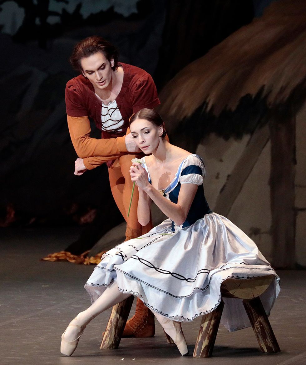 Smirnova sits on a wooden bench onstage in a blue dress holding a daisy with Belyakov leaning over her.