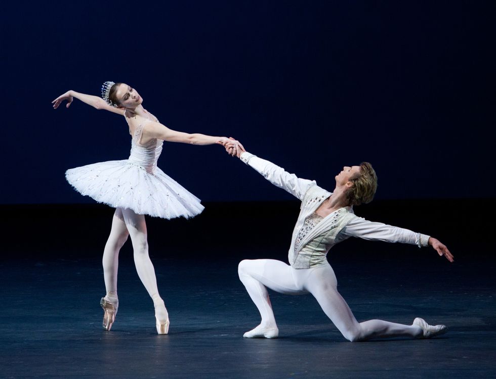 Smirnova, in a white tutu and crown, stands on pointe reaching away while also holding hands with her partner, who kneels on the floor.