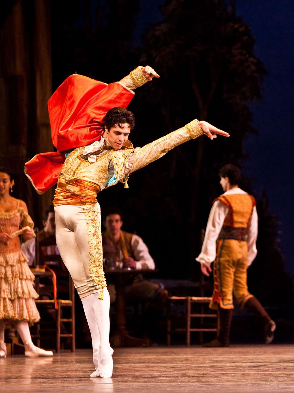 ALexandre Hammoudi, wearing white and gold tights, gold bolero jacket and red cape, stands in sixth position and arches over to his left, arms raised high and fingers pointed.