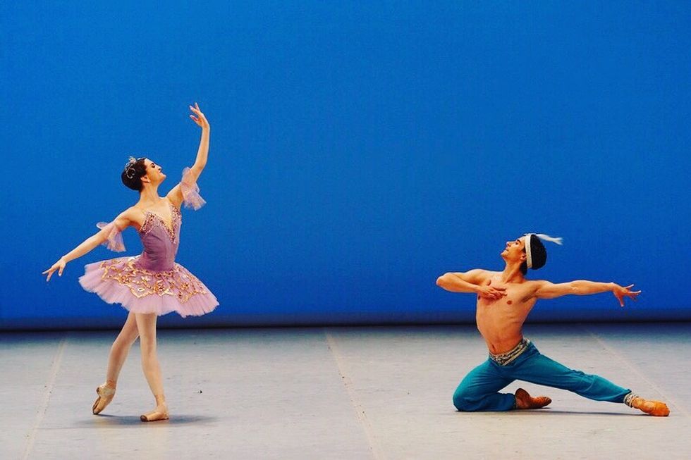 Joy Womack, in a purple tutu, stands in tendu derriere crois\u00e9 on the left looking up at her left hand. Mikhail Martynyuk, in blue harem pants, kneels with his left leg extended on the right side, his right arm gesturing toward his chest.