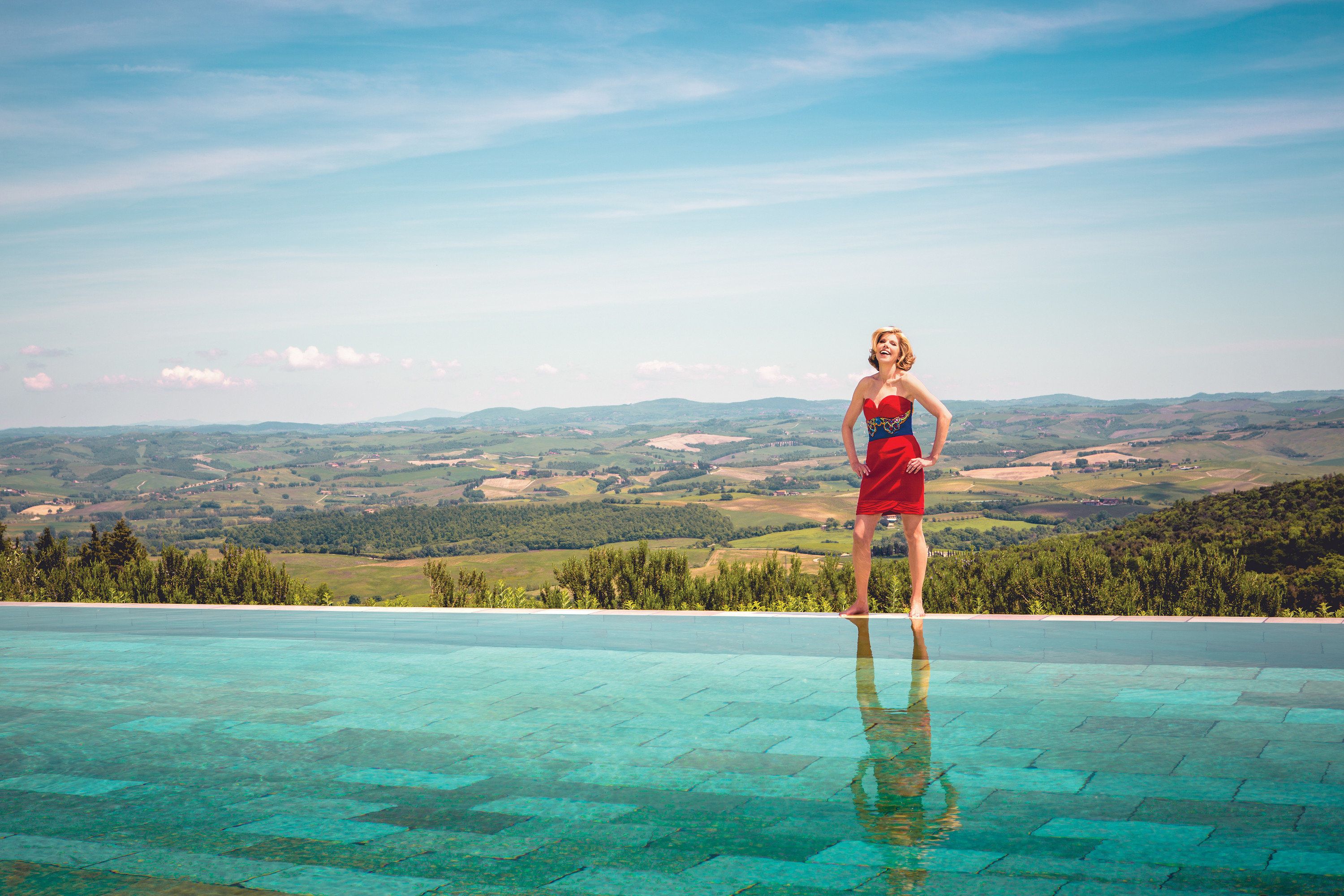 Christine Baranski stands poolside wearing a vibrant red and blue dress with a buccolic countryside stretching out behind her
