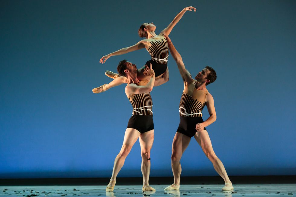 A male dancer props a woman dancer on his shoulder as she reaches up toward the sky and a third male dancer holds her left shoulder . All wear costumes of black brief bottoms and black-and-white striped tops.