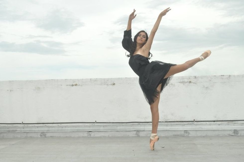 A young woman with long, dark hair and wearing a black dress, poses on a rooftop in piqu\u00e9 attitude, her arms high in a v-shape.