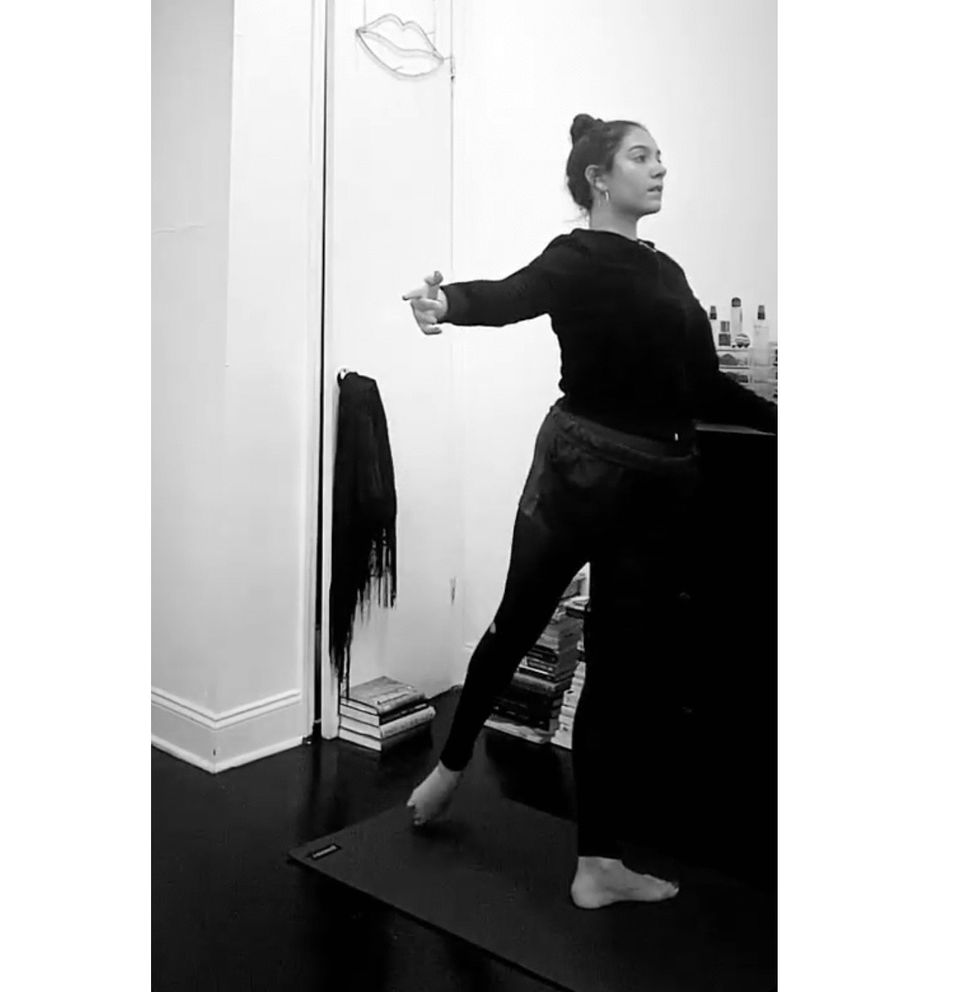 A young woman with her hair in a bun and wearing dark dance clothes holds onto a bedroom dresser and performs tendu derri\u00e9re.