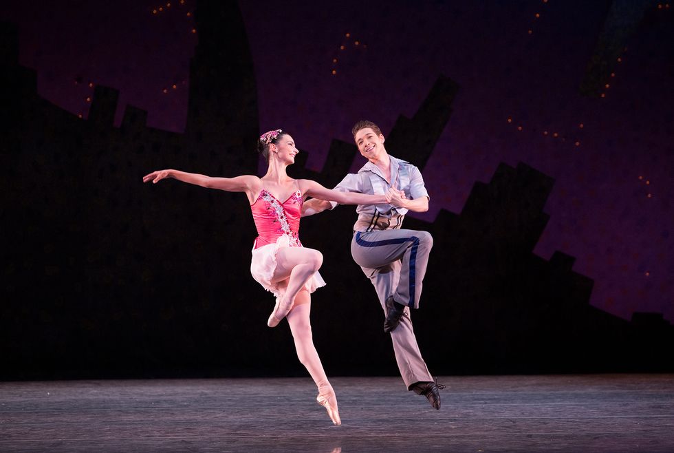 A ballerina in a pink sparkly dance dress and a male dancer in gray pants and a silver button-down shirt, hold hands and jump in pass\u00e9.