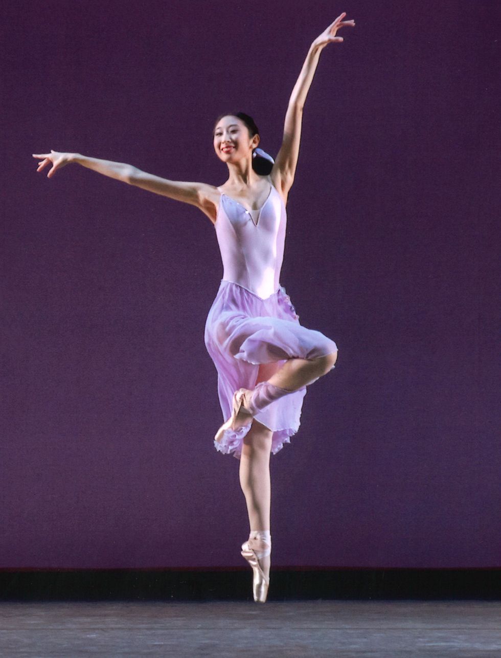 Lara Tong, in a purple dance dress, jumps with her right leg in pass\u00e9 and her left arm high in front of a purple stage backdrop.