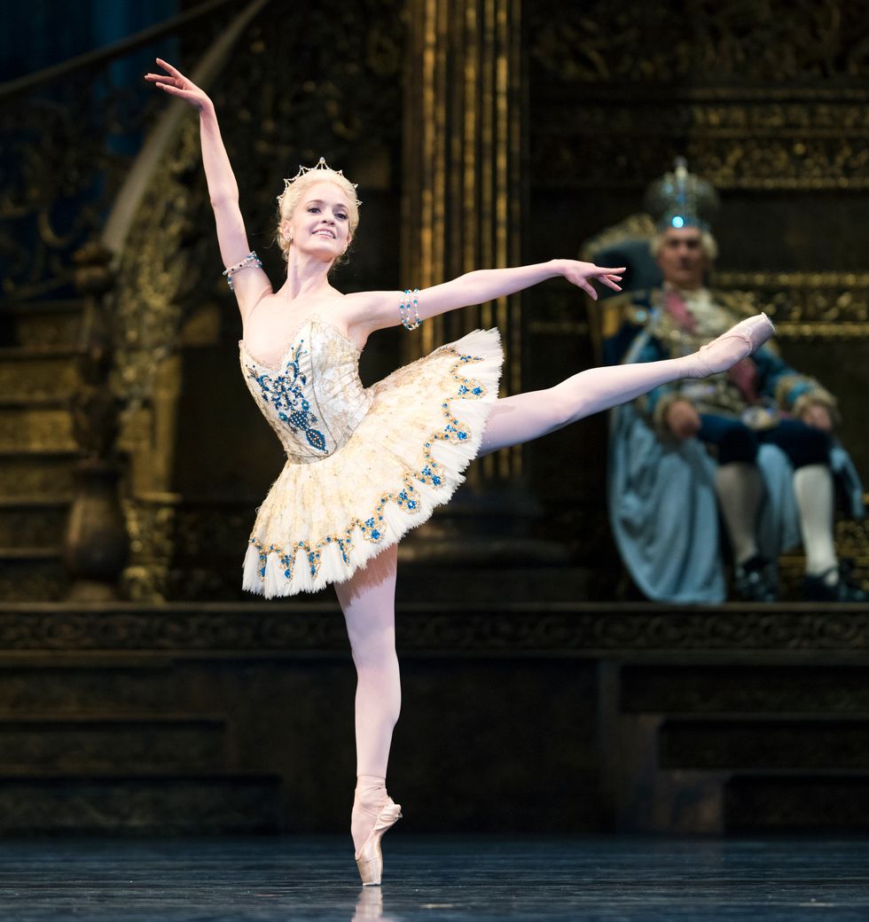 A ballerina in a white, gold and blue embellished tutu poses in first arabesque on pointe.