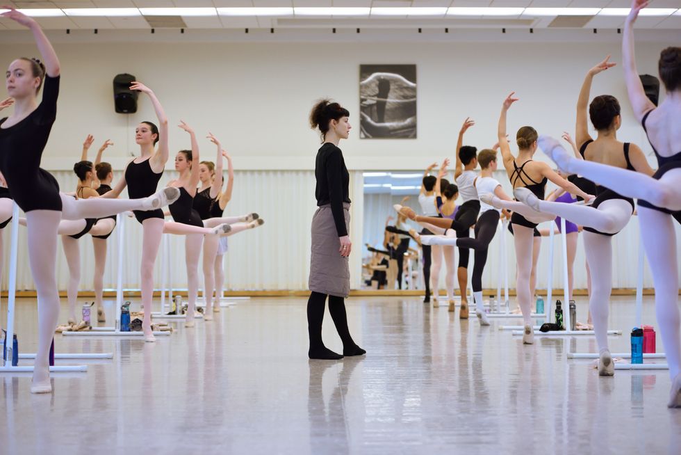 Wearing black tights and shirt and a brown skirt, Sofiane Sylve stands in between two barres of ballet students, who pose in attitude back.