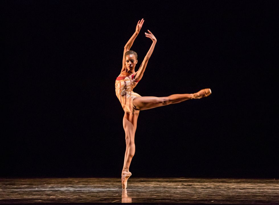 Wearing a beige and brown striped leotard with red embellishments, Dara Holmes does relev\u00e9 arabesque on her right leg and lifts both arms high into fifth position.