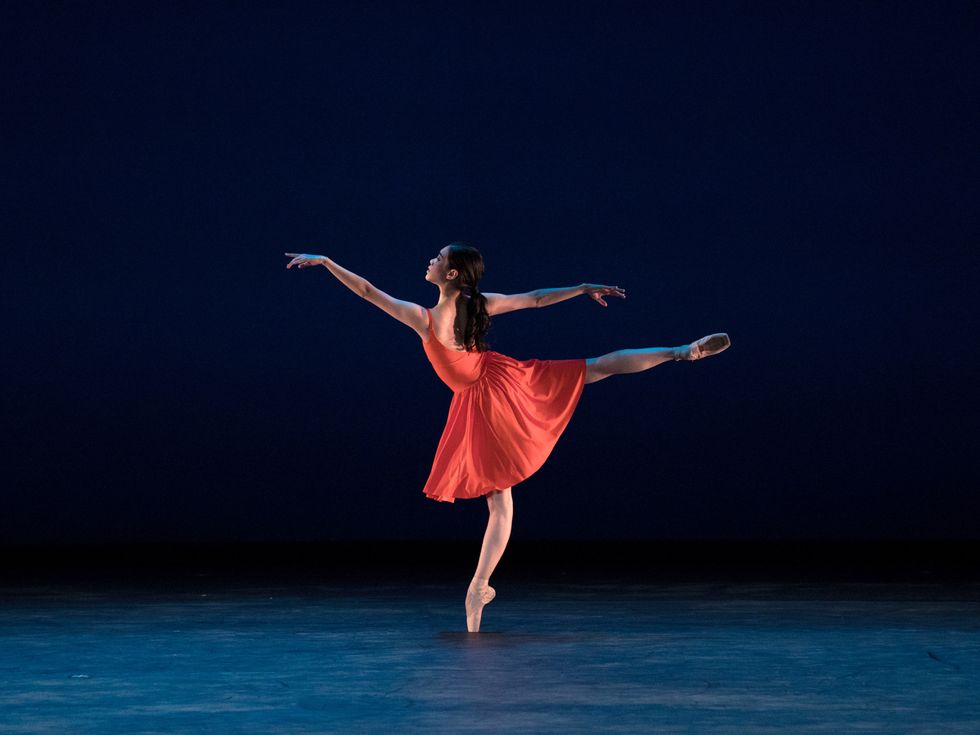 On a blue lit stage, Wanyue Qiao wears a red dance dress and does a piqu\u00e9 in second arabesque.