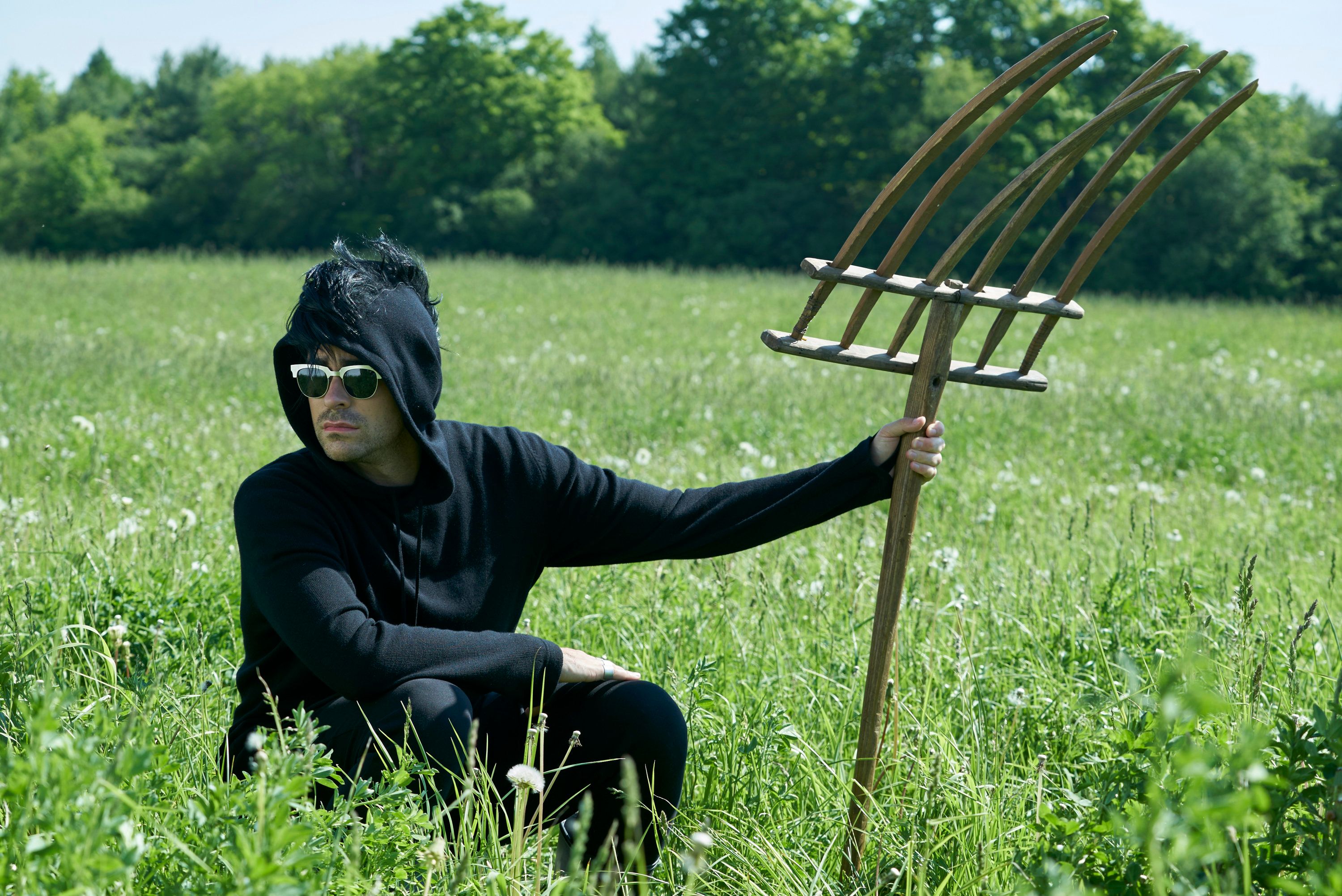 David crouches in a field holding a pitchfork and wearing a black hoodie with faux hawk