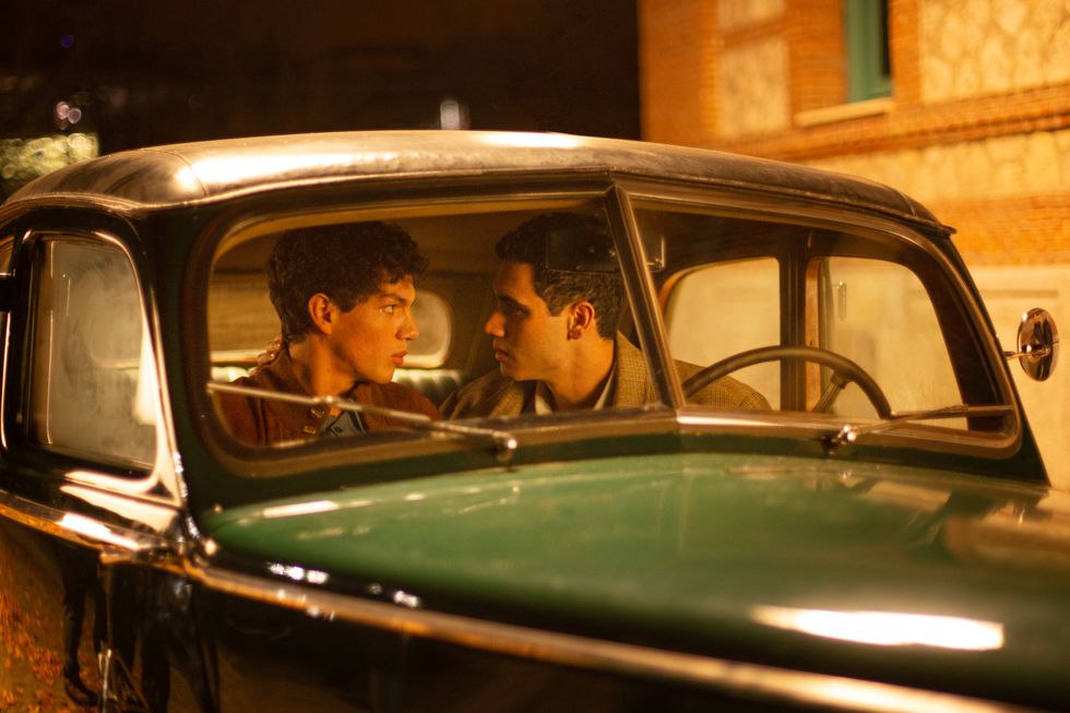Two men in brown jackets are shown talking closely in the front seat of a green 1950s-style car.