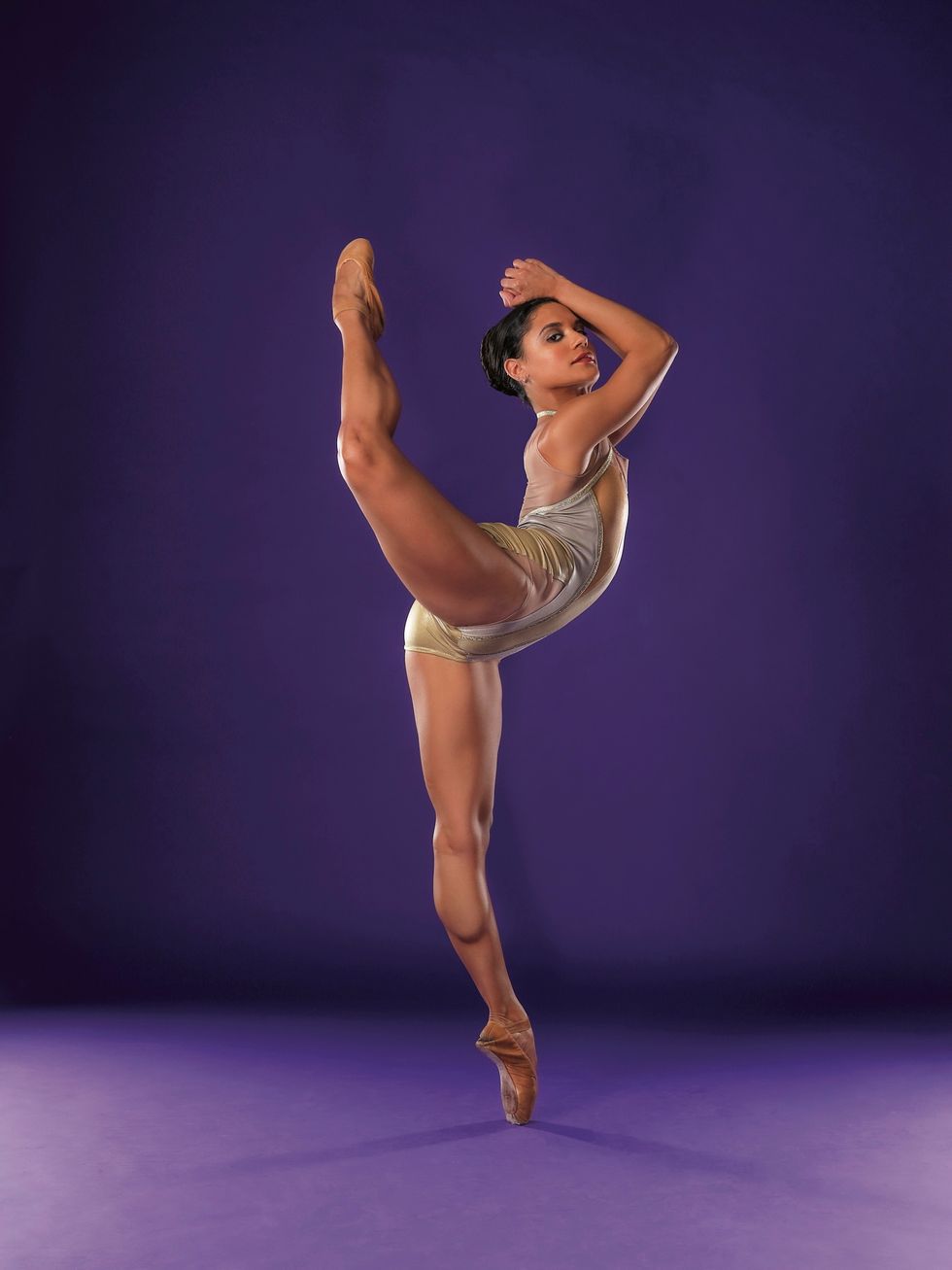 Tatiana Melendez, wearing a shiny gold leotard, stands on her left leg on pointe and kicks her bent right leg head height behnd her.