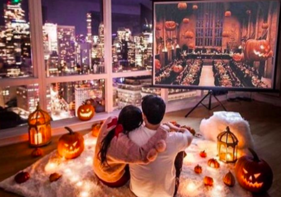 two people sitting on white blanket cuddling with pumpkins and lanterns around them and halloween movie on projector screen in front of them