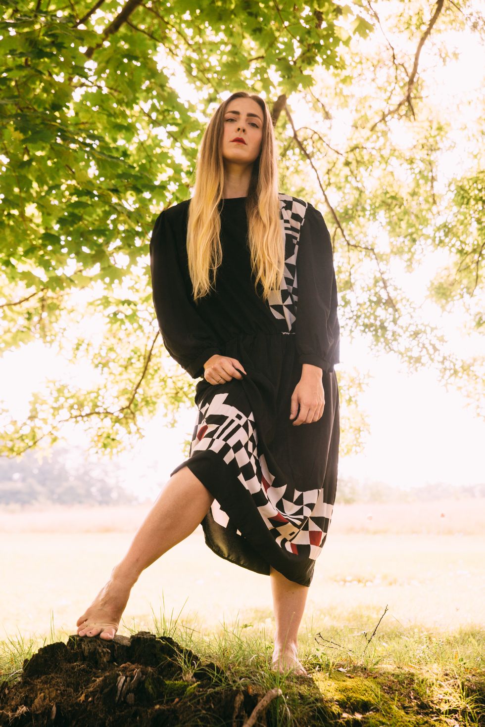 Jenn Freeman stands staring at the camera with one bare foot on a tree stump
