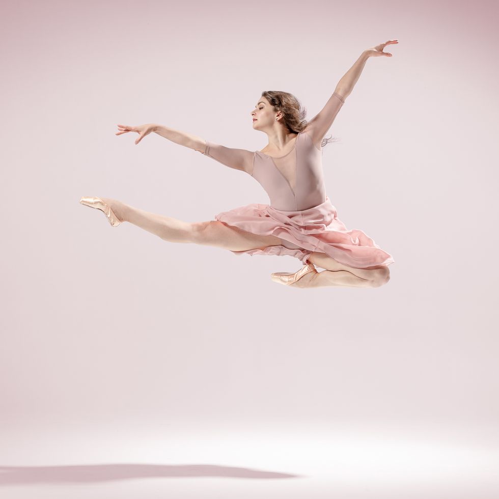 Kathryn Morgan, wearing a pink long-sleeved leotard and skirt, does a high Italian pas de chat in front of a pink backdrop.