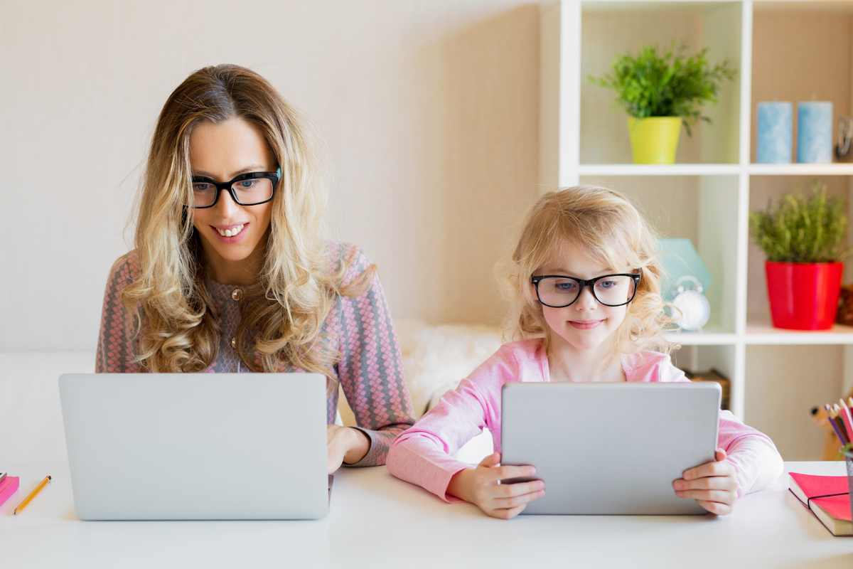 blonde woman and daughter in matching black glasses look at their laptops together at a desk