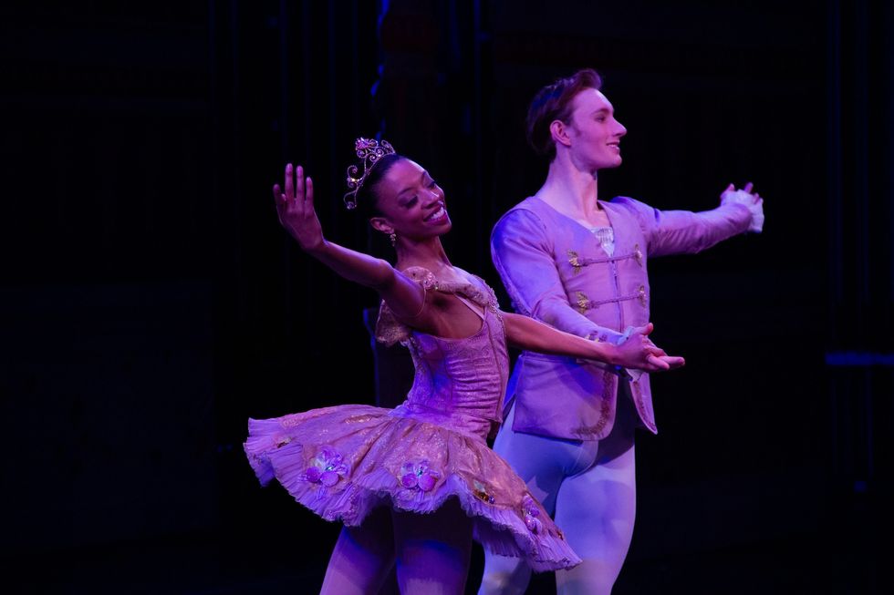 Whitney Huell, wearing a pink tutu and tiara, holds her male partner's right hand, while they both gesture out with their outside arms.