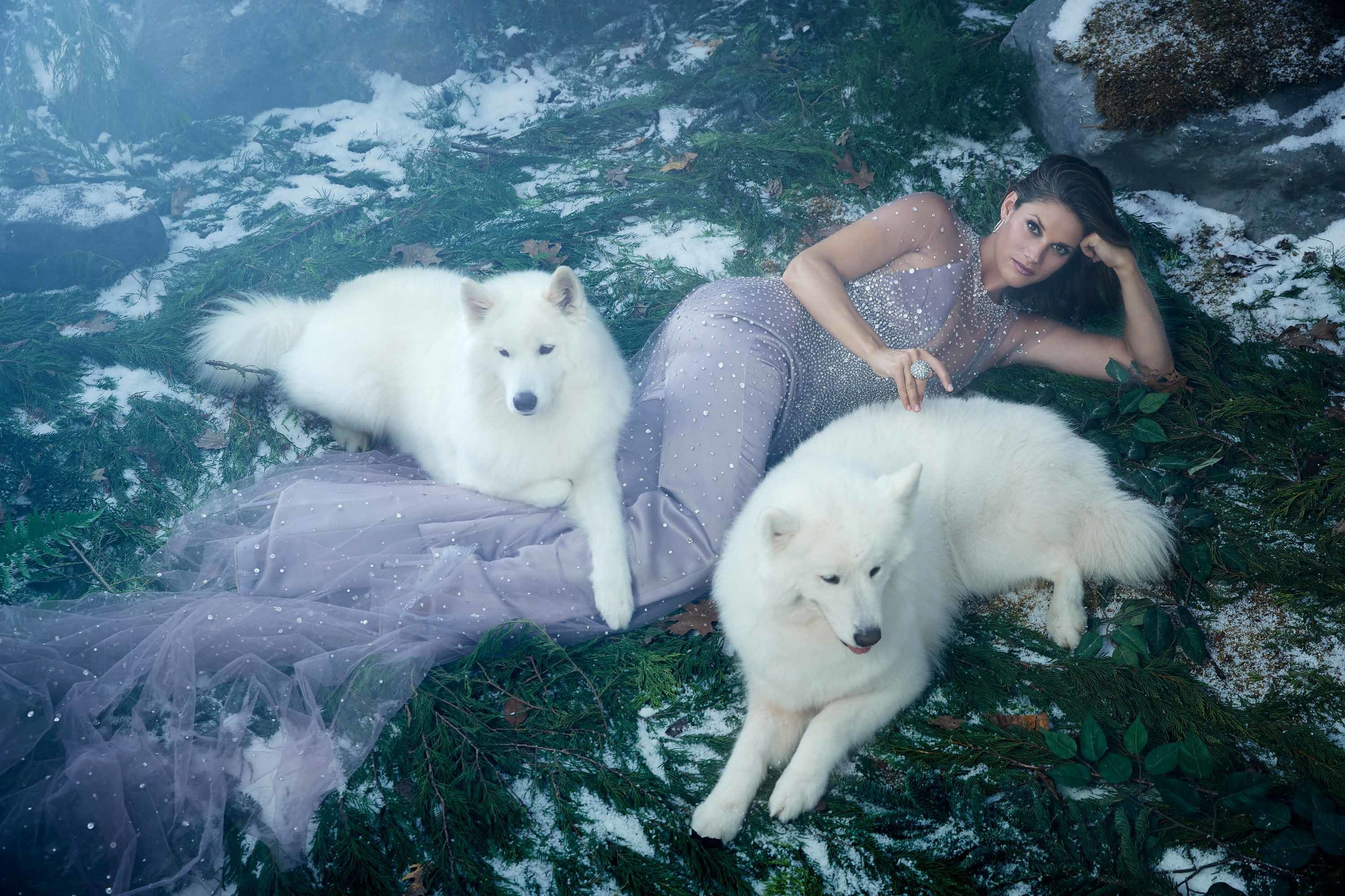 Actress Missy Peregrym of the show FBI wears a sheer and flowing dress while reclining on the ground with two white dogs.