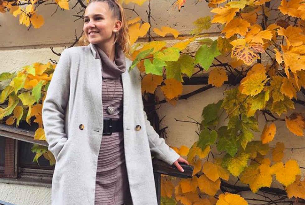person standing in front of trees with orange leaves wearing dress and pea coat
