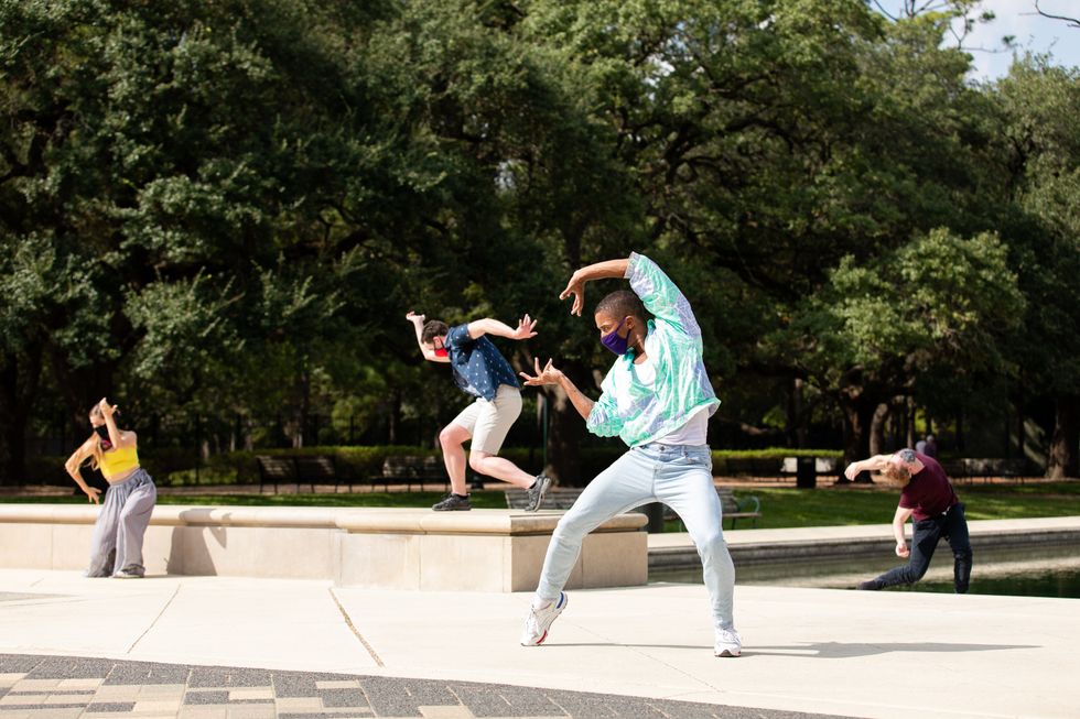 A young male dancer in jeans and an aqua jacket stands outside in a parallel second position pli\u00e9, pops his right foot and curves his left arm overhead. A female and two male dancers in street clothes dance behind him.
