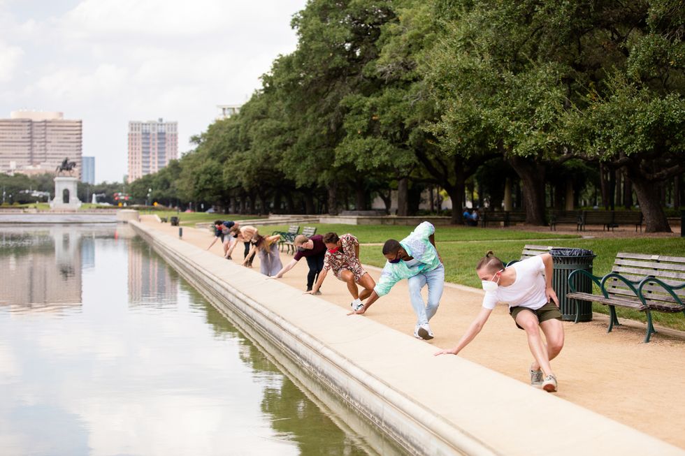 A line of male and female dancers in street clothes stand in a line and reach down to touch the side of a reflecting pool in a city park.