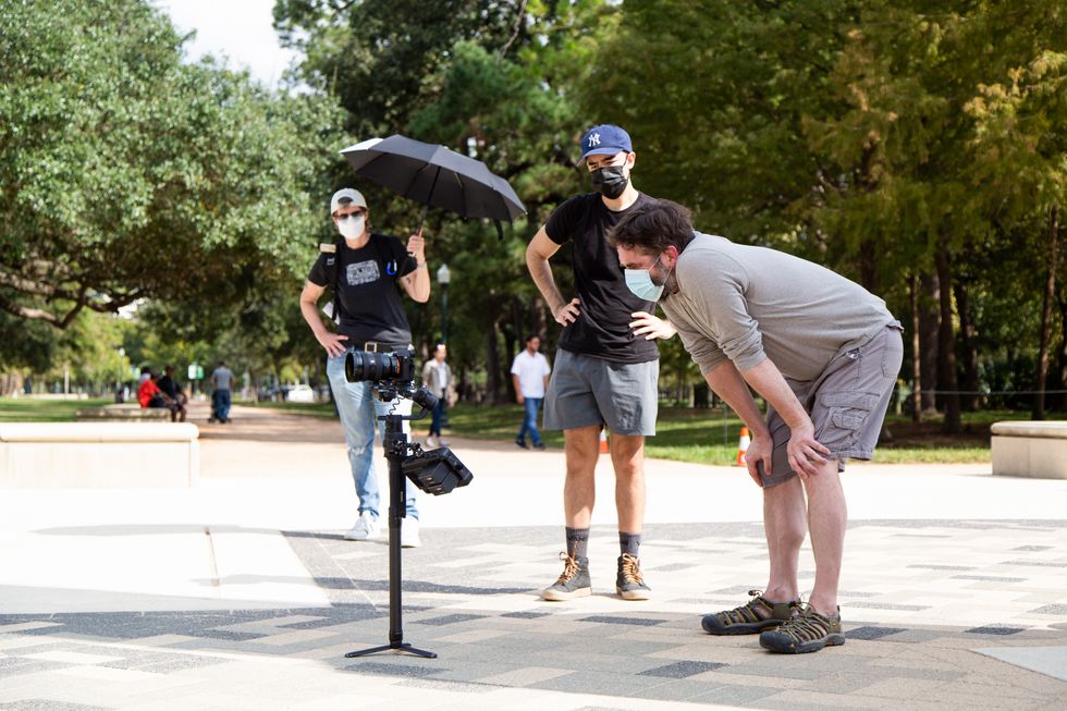 A man in a gray long-sleeved T-shirt and gray shorts leans down with his hands on his knees and looks into a camera on a tripod. Two men in black T-shirts stand behind him in the park.