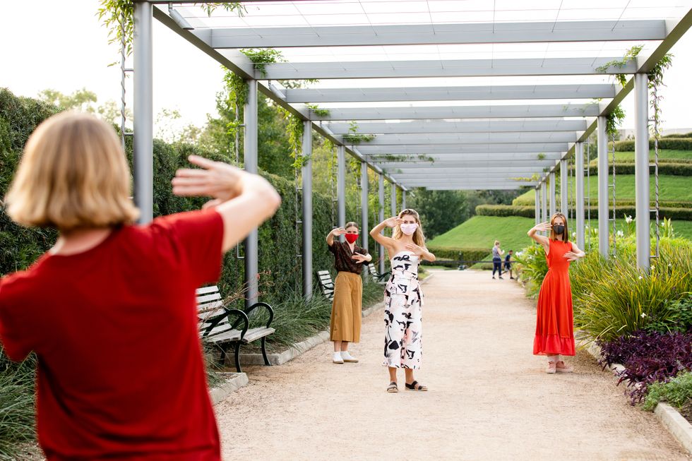 At a city park, a woman in a red shirt stands in the foreground and makes a pushing motion with her hands. Three female dancers in street clothes stand in a triangle formation in front of her and copy her.