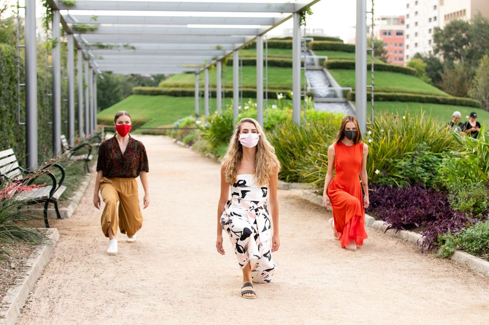 Three women dancers in brightly colored street clothes crouch down onto the gravel on their left knee, their arms hanging by their sides. They are in a city park surrounded by plants and stand under a metal canopy.