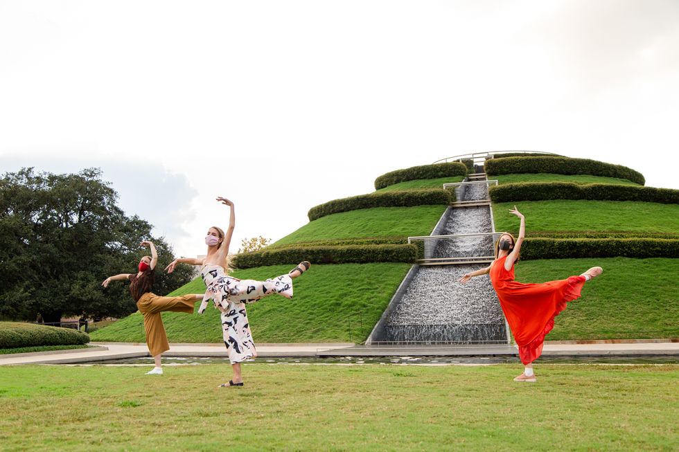 Three female dancers in brightly colored street clothes pose in attitude derriere in front of a manicured hilltop fountain.