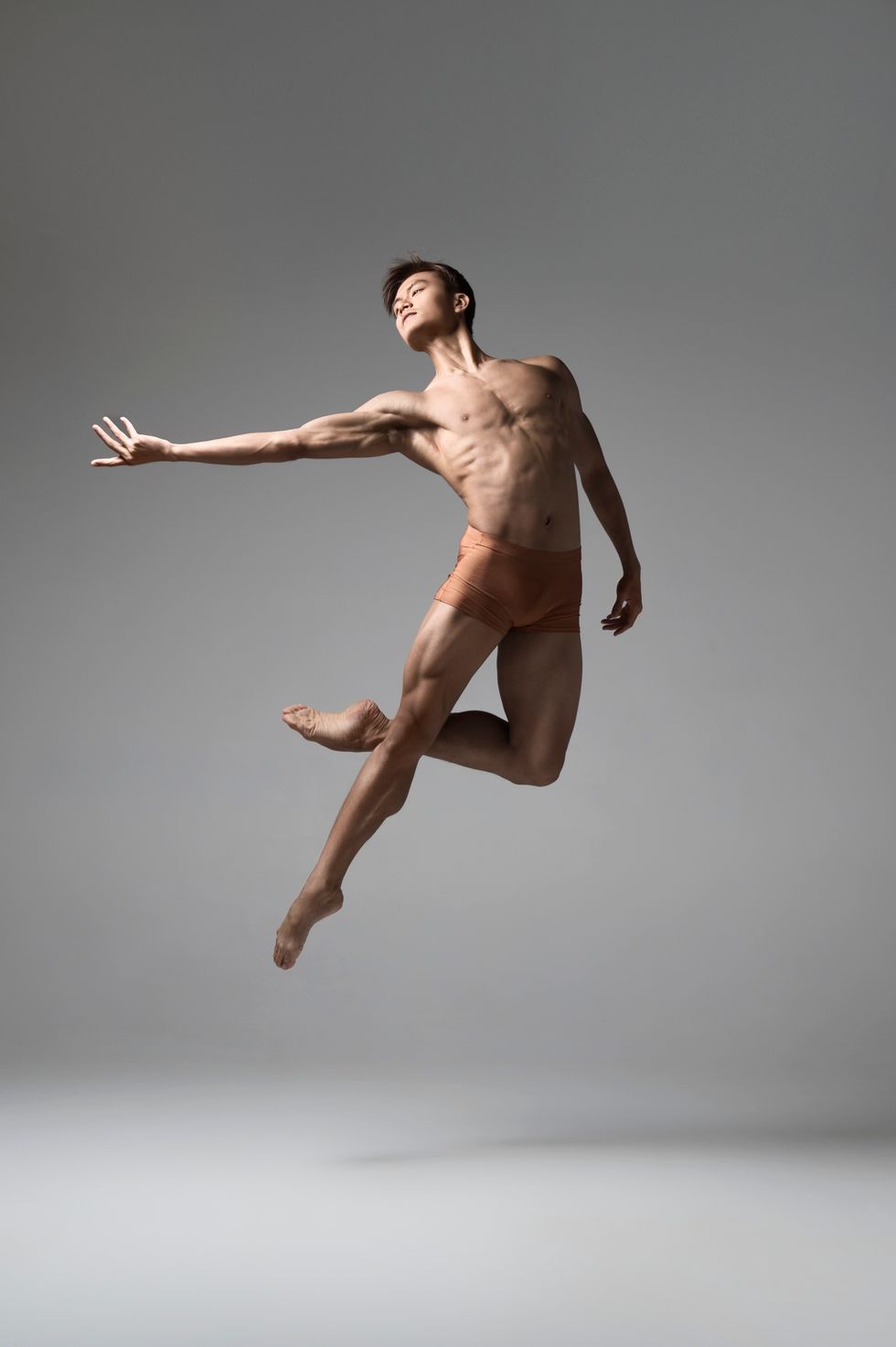Wearing tan booty shorts, Chun Wai Chan jumps into a saut\u00e9 with his left leg in pass\u00e9, extending his right arm out to the side.