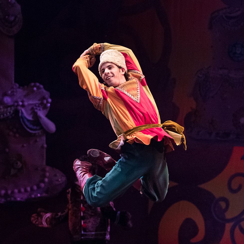 Taylor Carrasco, in a colorful Russian folk costume , white furry hat and purple boots, arches his back and jumps with his legs bent back behind him and his arms clasped over his head.