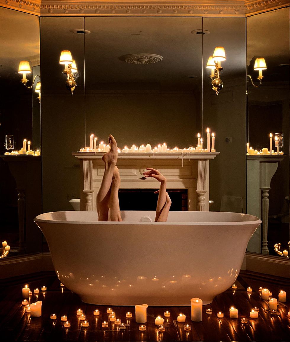 Four limbs delicately peek out over the top of a bathtub surrounded by candles. 
