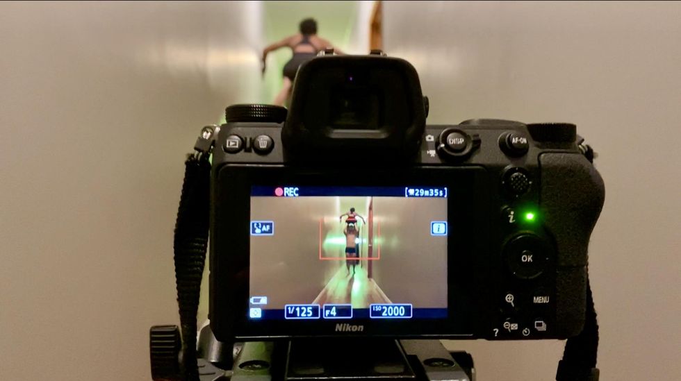 A camera with a green light captures two dancers on top of each other in a long white hallway. The top of one peeks out above the camera in the distance.