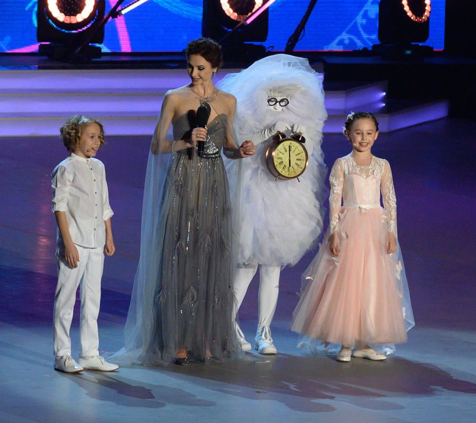 Two children stand on either side of a be-gowned Svetlana Zakharova. A character in white with a clock stands at her side as well.