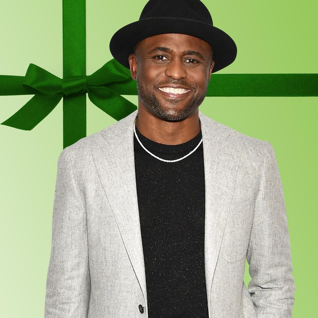Actor Wayne Brady smioles while wearing a light grey suit and black hat.