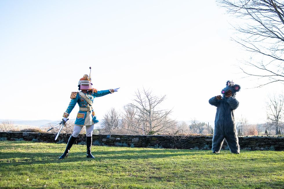 A male dancer in a Nutcracker costume and mask stands with his weight on his left leg and points towards another dancer in a plush Mouse King costume. They stand outside on a grassy lawn and in front of a low stone fence.