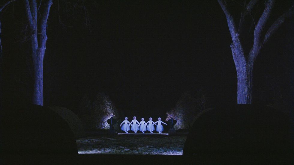 A group of five female dancers in long white tutus and white crowns stand in line in B+ at nightfall on an outdoor stage. Two large trees flank the stage.