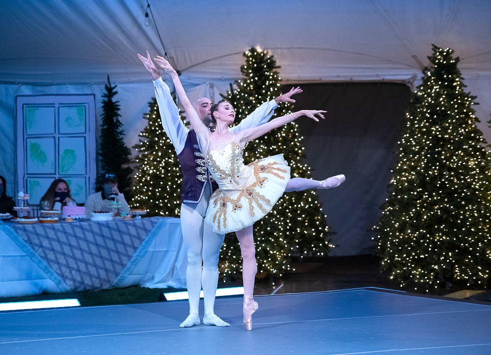 A female dander in a white and gold tutu and pink tights does piqu\u00e9 first arabesque on pointe on her right leg. Her partner, in white tights and a purple tunic, stands directly behind her with his arms held high. Three audience members sit at a table behind them, while three Christmas trees glitter to the right of them.