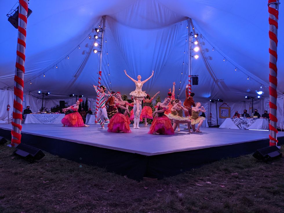 A large group of dancers in brightly colored costumes pose in a tableau, with several on their knee and the main ballerina lifted high in the air by her partner.
