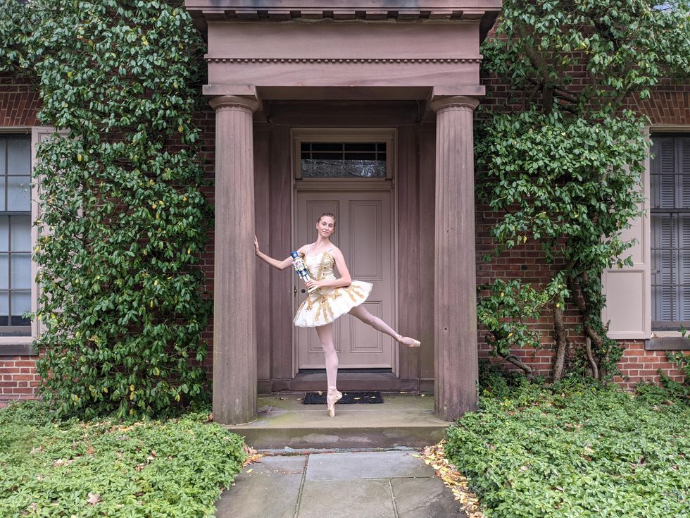 A white ballerina wearing a white and gold tutu, pink tights and pointe shoes poses in a low arabesque on the front stoop of a large brick estate covered in ivy. She holds a nutcracker doll in her left hand and rests her right hand on a large pillar for balance.