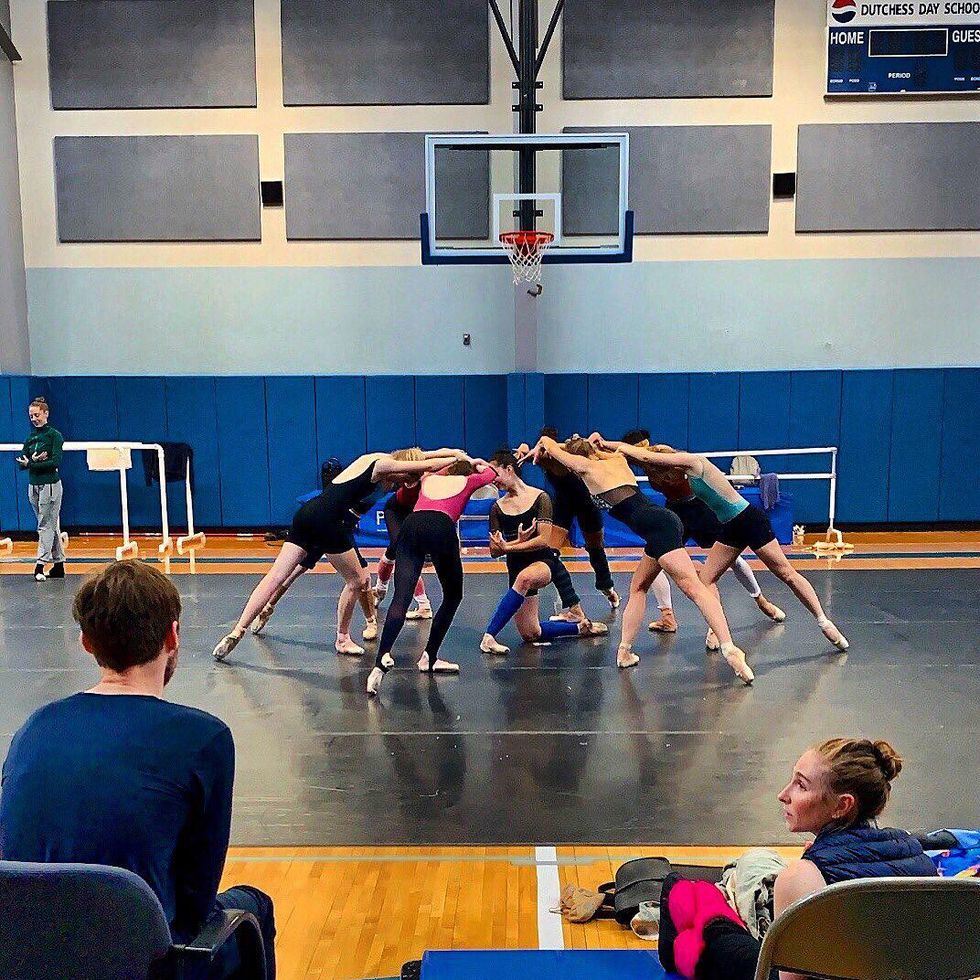 A group of female dancers wearing practice clothes rehearse in a school gym. Eight of them stand in tendu derriere and bend their bodies forward, encircling another dancer posed on her right knee on the floor. A man and woman sit in chairs in front of them, watching from a distance.