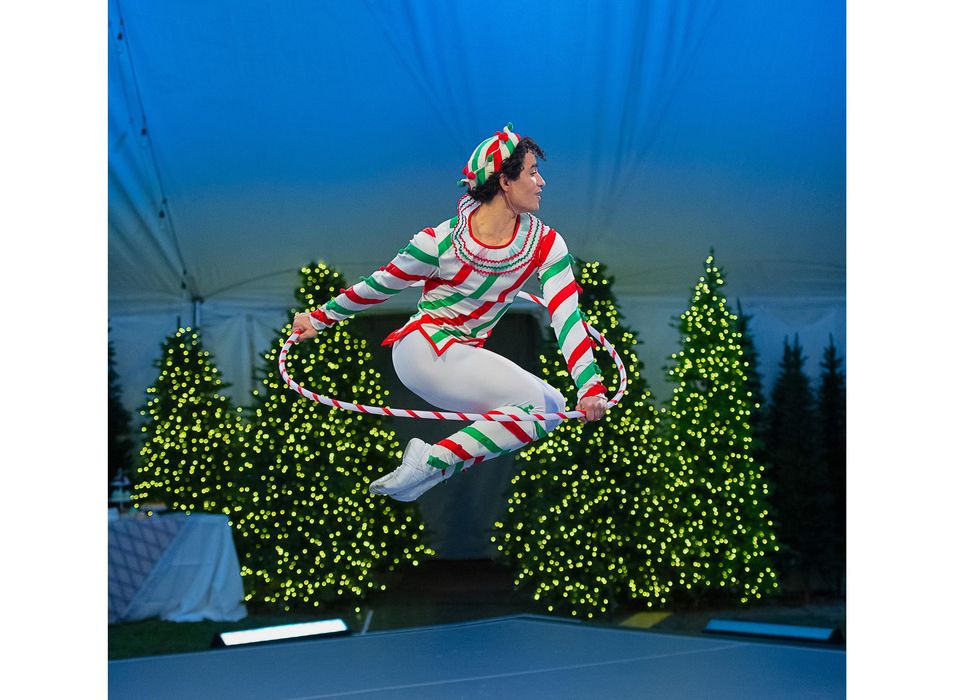 A male dancer in a green, white and red striped tunic, tights and hat holds a hula hoop and jumps up, tucking his legs underneath him.