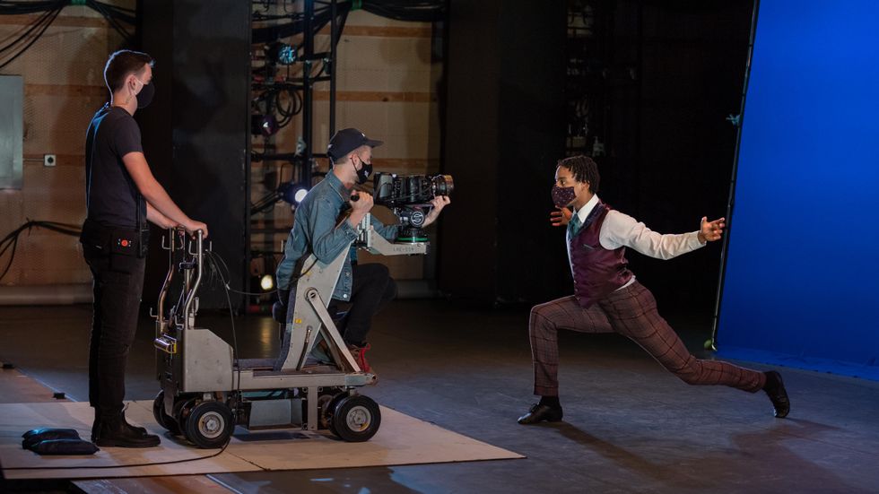 A Black dancer in a black facemask, brown pants, white shirt, brown vest and green tie lunges towards a movie camera with his arms held out wide.