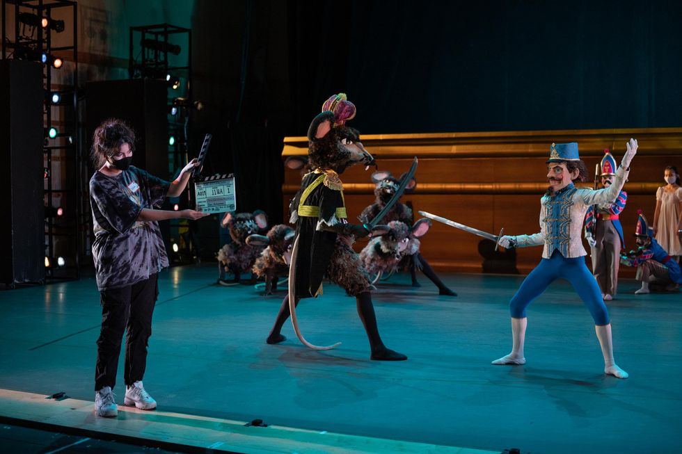 A woman in a gray top and black pants holds a clapperboard in front two dancers on a green-lit stage. The dancer on the left wears a rat costume with a black and yellow jacket, while the dancer on the right wears a Nutcracker costume made of blue leggins over white tights, a white, gold and blue jacket, and large Nutcracker head.