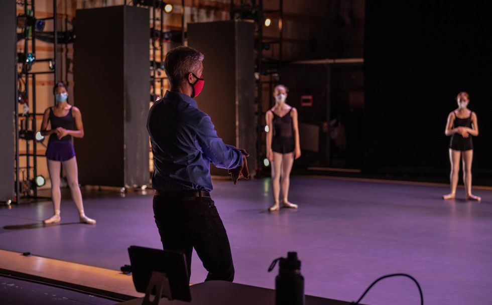 A gray-haired man in a red facemask, blue shirt and black pants stands in front of a stage and addresses three female dancers in pink tights, pointe shoes and black loetards.