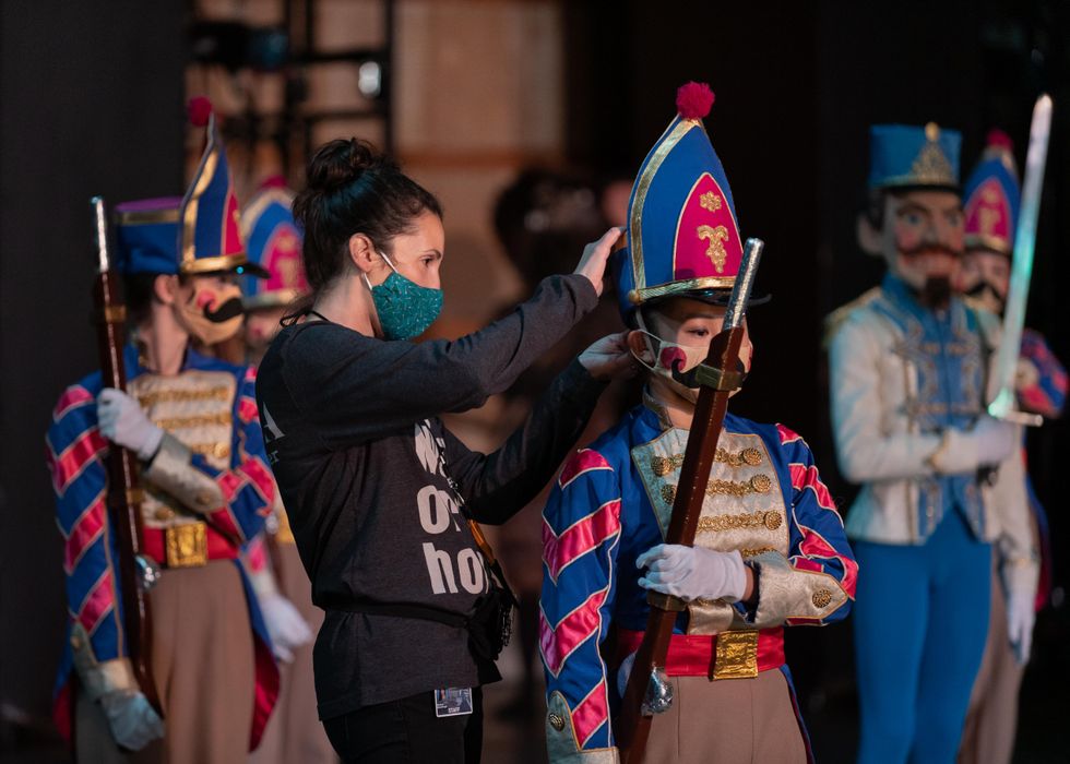 A woman in a black shirt and pants and green facemask adjusts the gat on a young dancer dressed as a toy soldier. The dancer's hat and jacket are blue and fuchsia, and she holds a brown shotgun prop down by her right side.