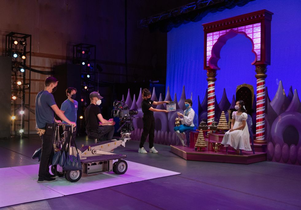 A film crew of three men stand around a movie camera on a purple-let stage as a woman in black holds a clapboard. In front of them two dancers costumed as a prince and Clara from The Nutcracker sit on a set-piece with candy cane pillars and tiny Christmas trees.