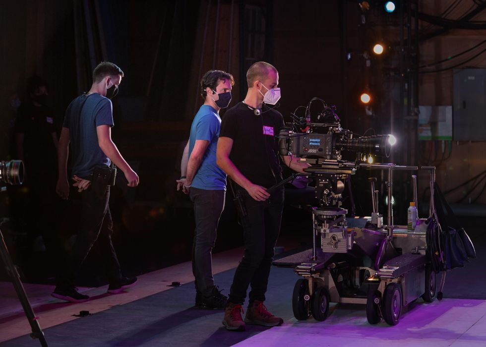 A white man in a blue T-shirt and black pants stands behind another white man in a black T-shirt and pants managing a large film camera. Another man behind them walks past towards the stage wings.